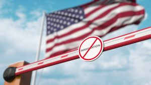 Closed boom barrier with stop immigration sign against the American flag. Border closure or immigration ban in the USA.Extra questioning at border 2026